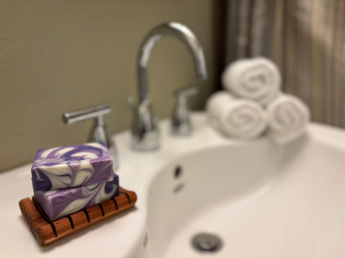 A close-up of a purple and white marbled soap bar resting on a small wooden soap dish on a bathroom sink, with a chrome faucet and rolled white towels blurred in the background.