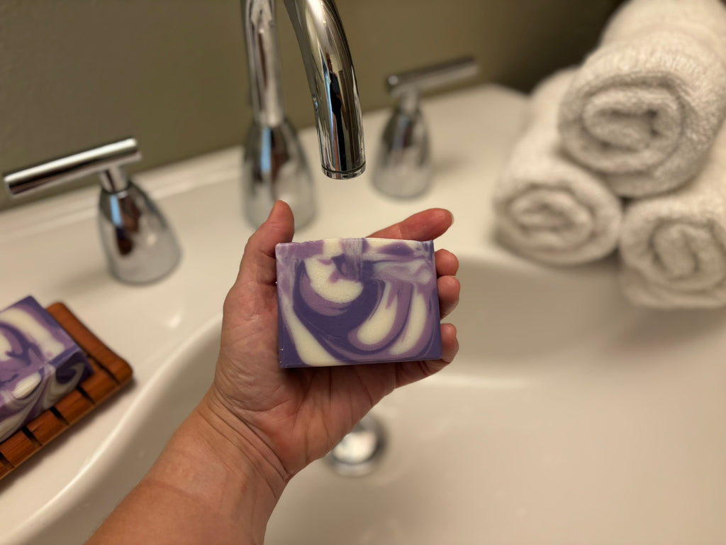 A hand holds a rectangular bar of purple and white marbled soap in front of a bathroom sink with a chrome faucet and rolled white towels in the background.