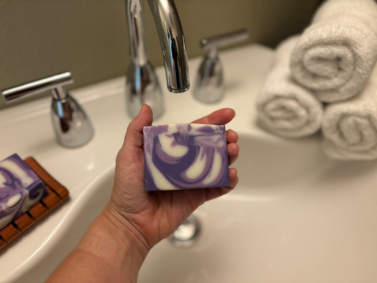 A hand holds a rectangular bar of purple and white marbled soap in front of a bathroom sink with a chrome faucet and rolled white towels in the background.