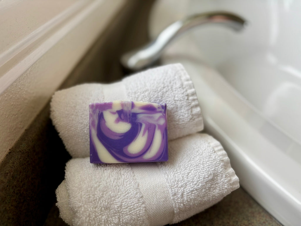 A rectangular bar of purple and white marbled soap rests on two folded white towels beside a white sink and faucet.