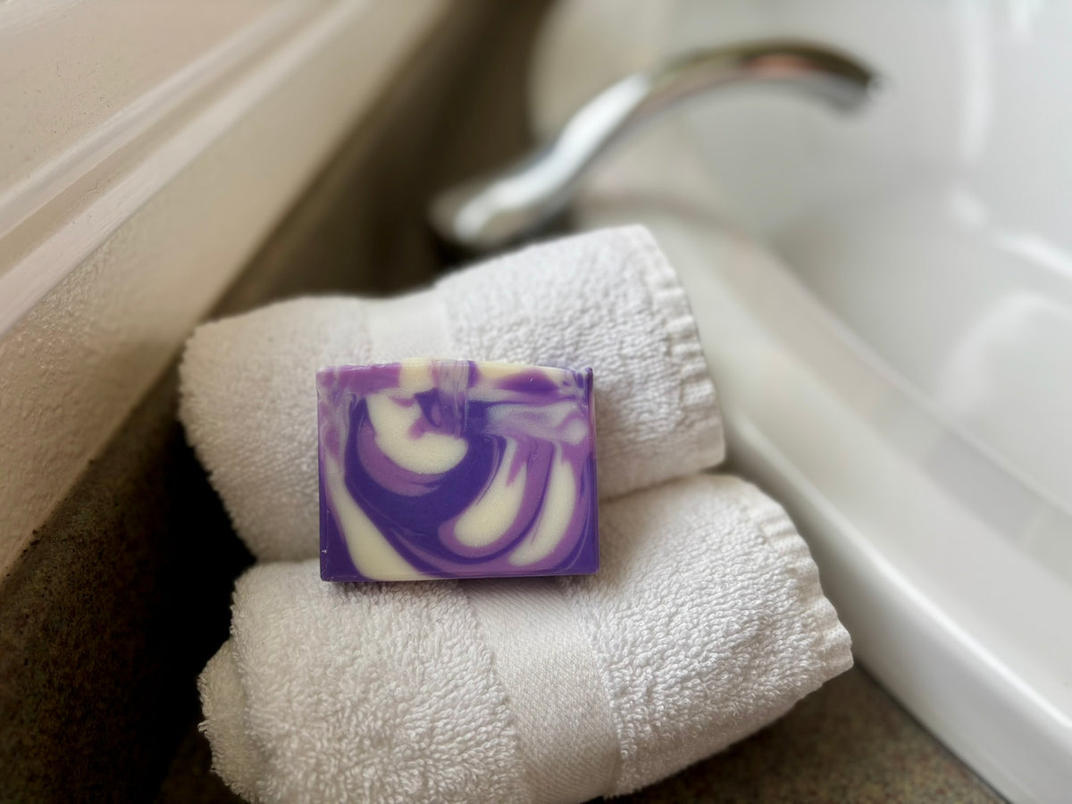A rectangular bar of purple and white marbled soap rests on two folded white towels beside a white sink and faucet.