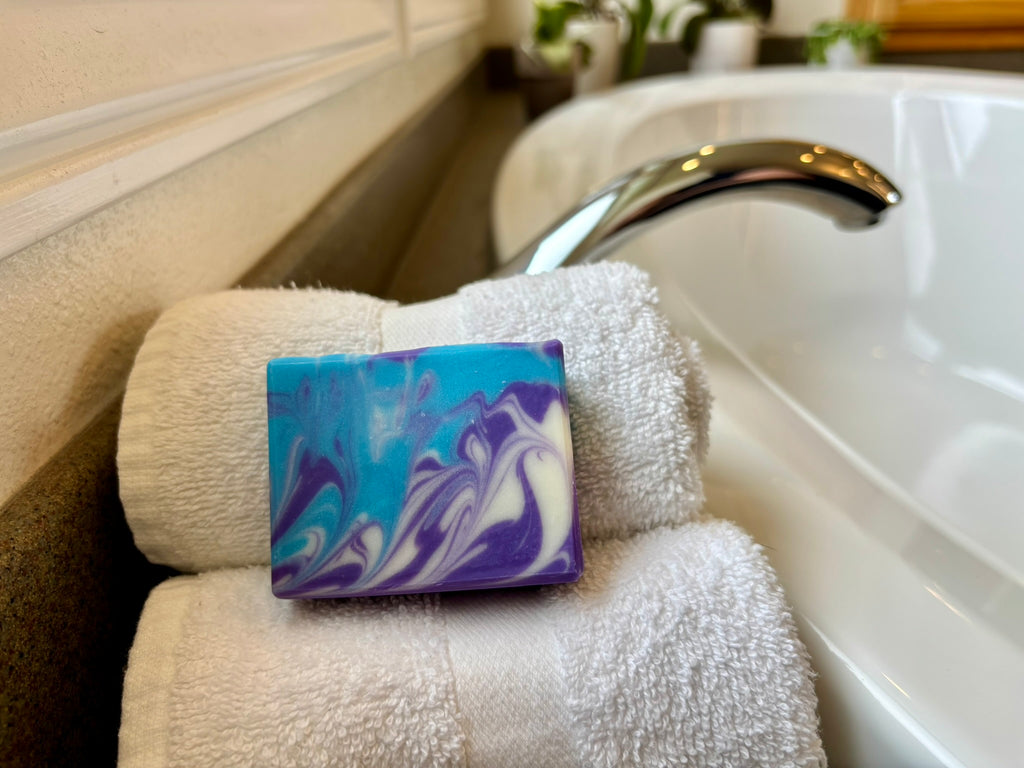 A rectangular bar of handmade soap with a blue, purple, and white marbled swirl pattern, resting on a stack of white towels next to a white bathtub faucet.