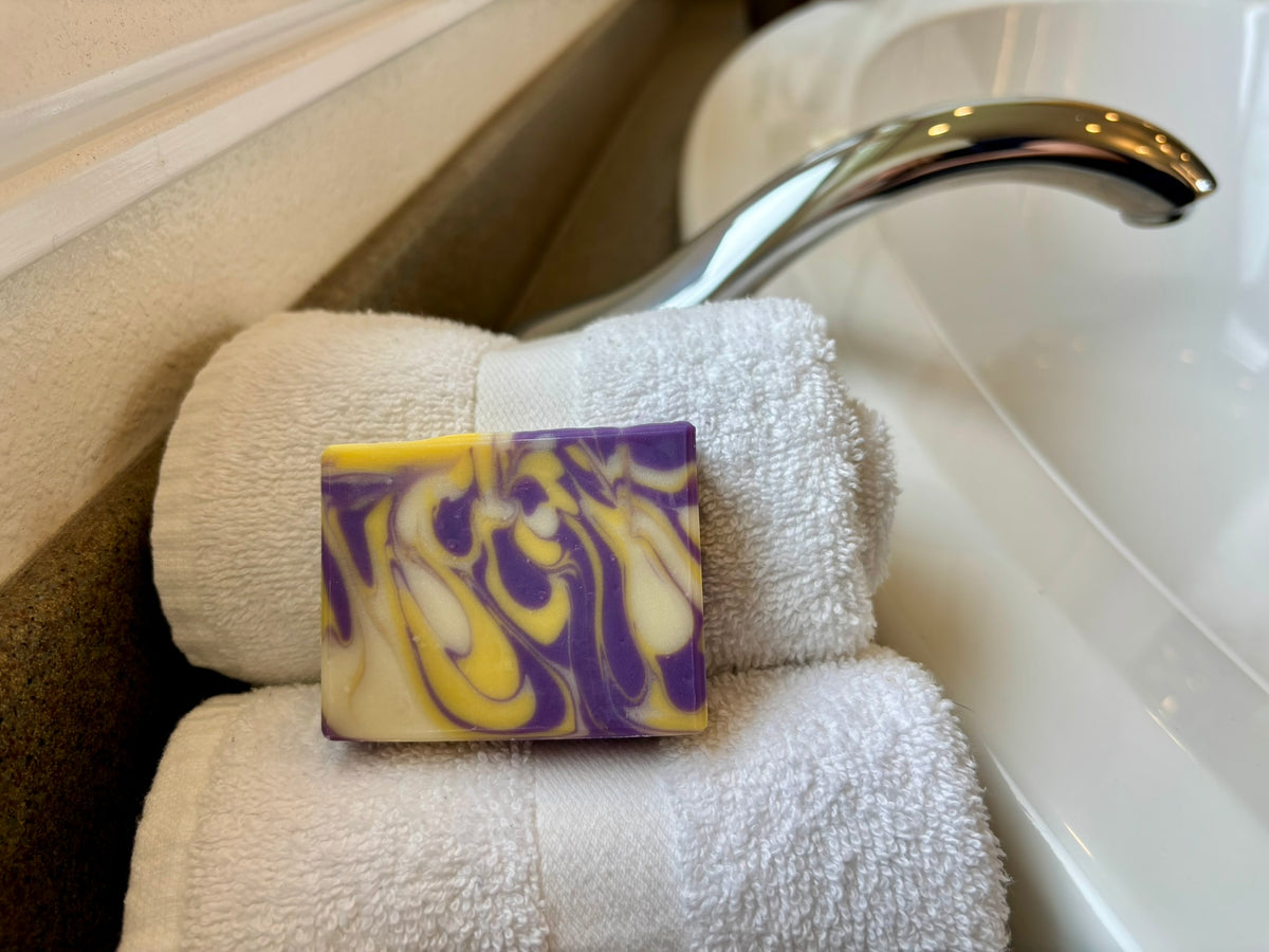 A rectangular bar of soap with a purple and yellow marbled swirl pattern, resting on a stack of white towels next to a chrome faucet.