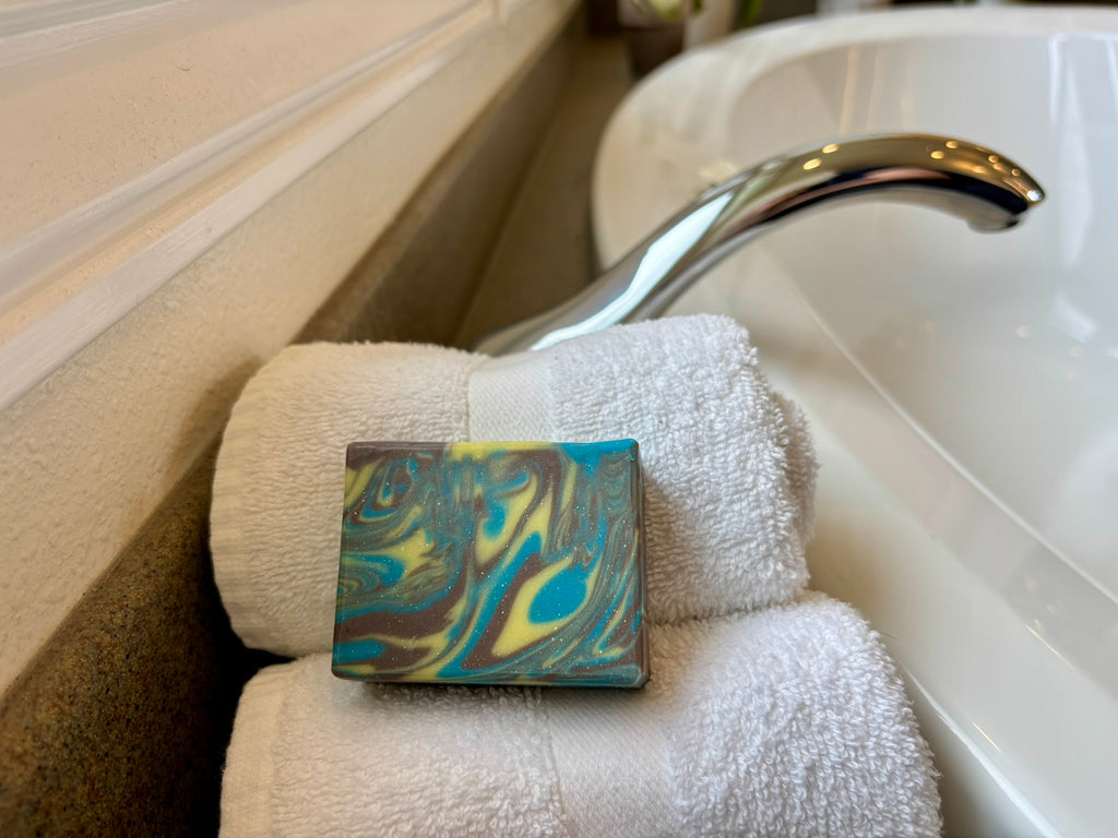 A rectangular bar of soap with a marbled blue, yellow, and brown pattern rests on folded white towels beside a chrome faucet and white tub.