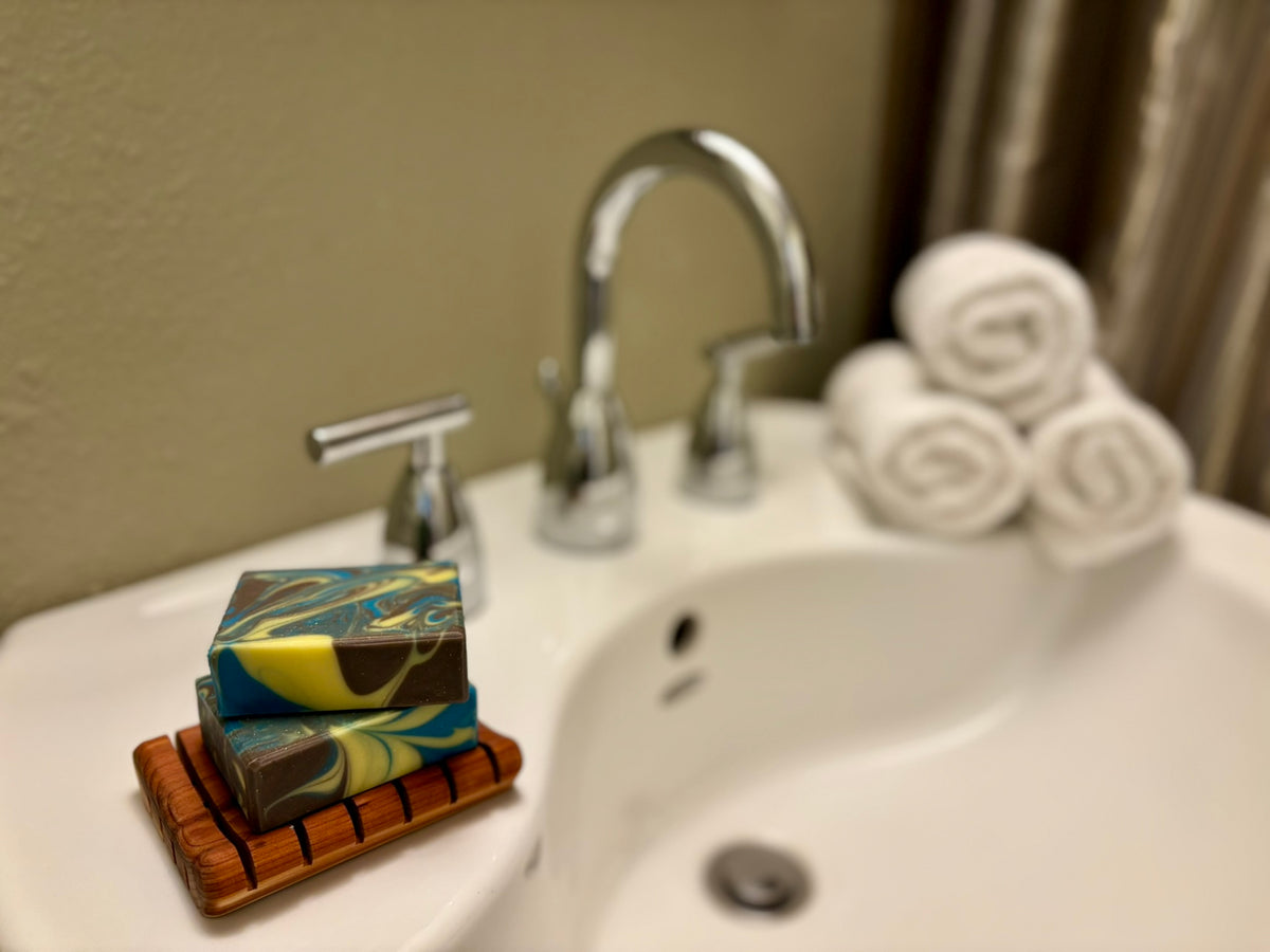 A stack of marbled soap bars in blue, yellow, and brown sits on a wooden soap dish on a white bathroom sink, with a chrome faucet, rolled towels, and a beige wall in the background.