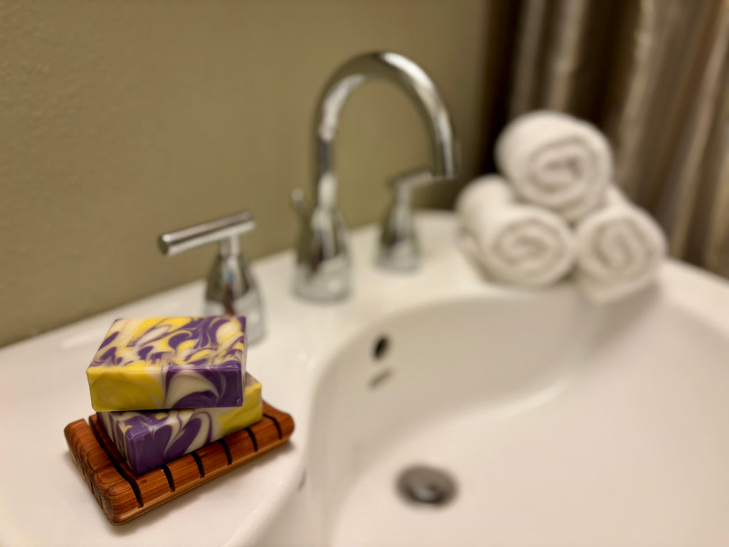 A stack of two marbled purple and yellow soap bars sits on a small wooden soap dish on a white bathroom sink, with a chrome faucet, rolled white towels, and a beige wall in the background.