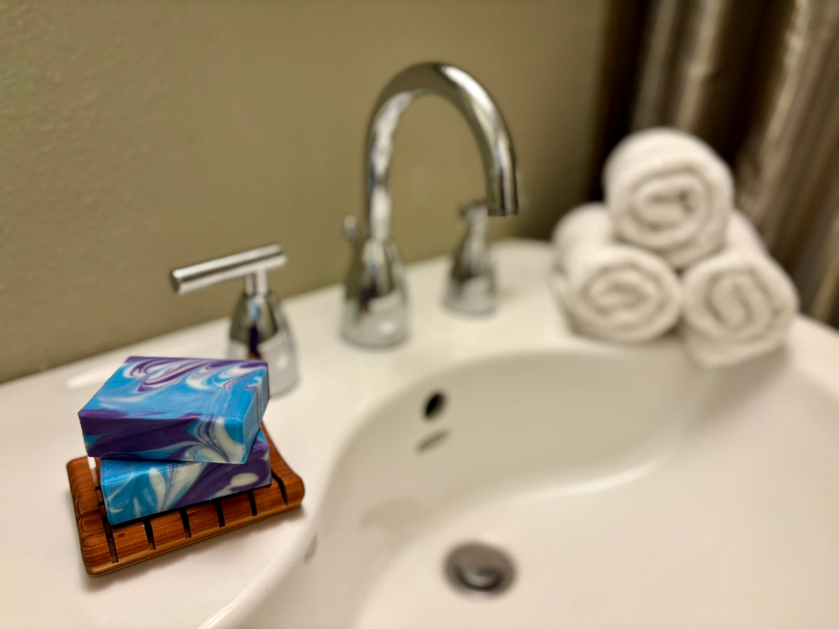 A close-up of a bathroom sink with a chrome faucet, a stack of blue and purple marbled soap bars on a small wooden dish, and three rolled white towels on the counter.