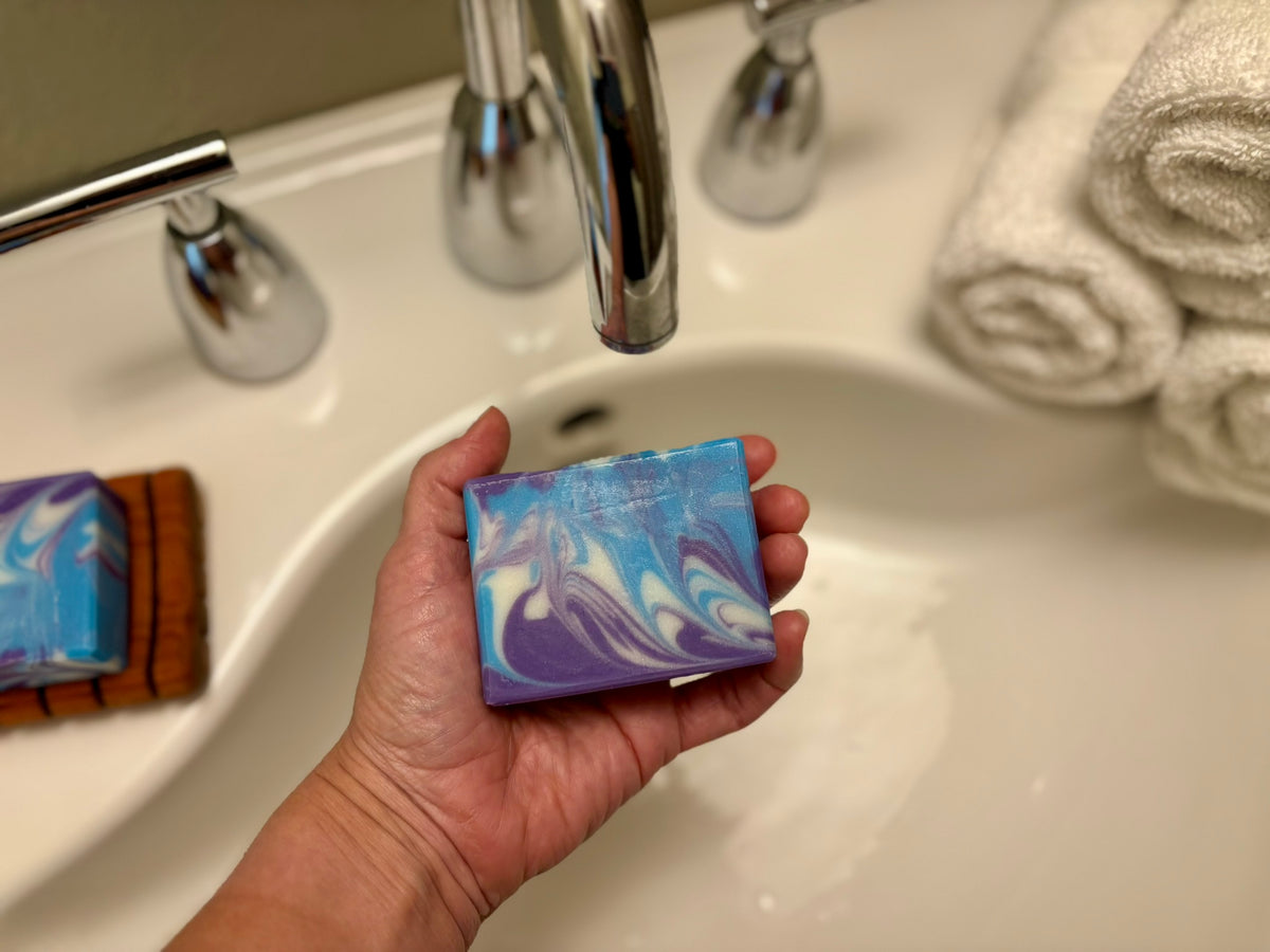 A hand holds a rectangular bar of soap with a blue, purple, and white marbled pattern in front of a bathroom sink with a chrome faucet. Another soap bar sits on the sink ledge to the left, and rolled white towels are visible in the background.