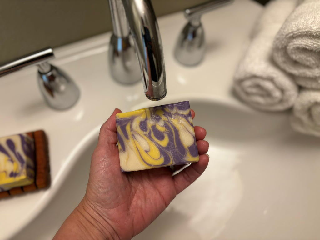 A hand holds a rectangular bar of marbled soap with purple, yellow, and cream swirls under a chrome faucet in a bathroom sink, with rolled towels visible in the background.
