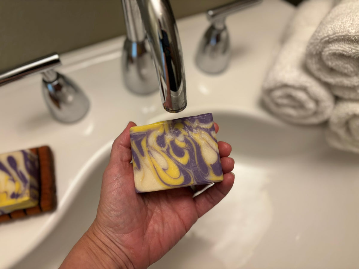 A hand holds a rectangular bar of marbled soap with purple, yellow, and cream swirls under a chrome faucet in a bathroom sink, with rolled towels visible in the background.