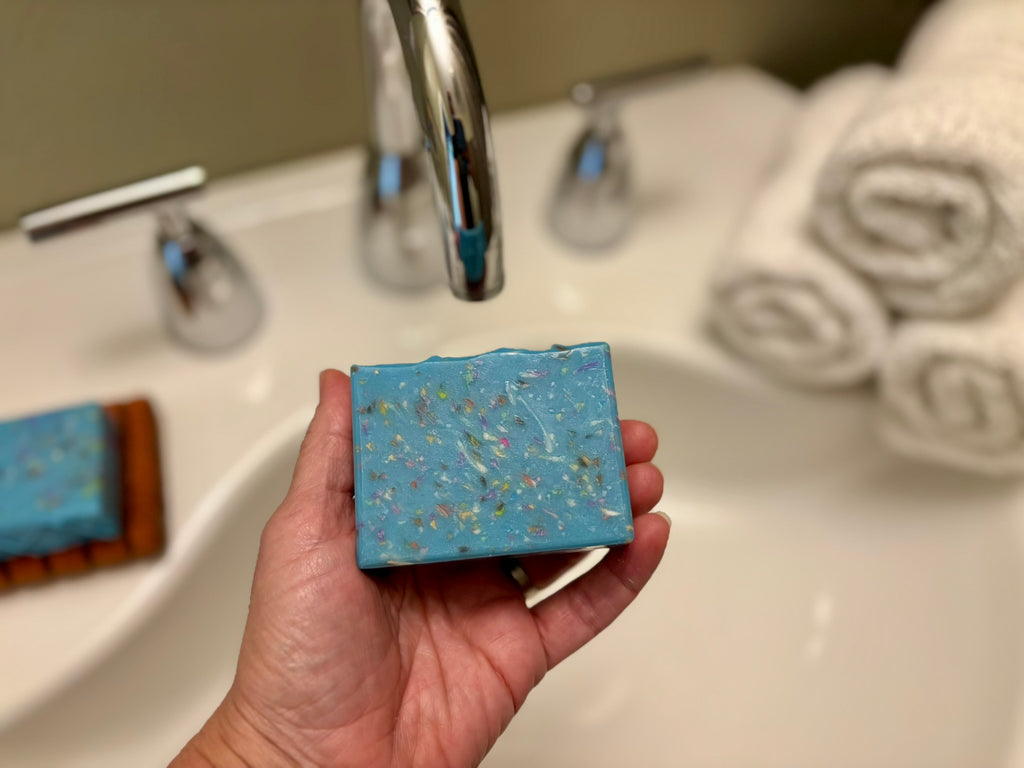 A hand holds a rectangular blue soap bar with colorful flecks under a chrome faucet in a bathroom sink, with rolled towels blurred in the background.