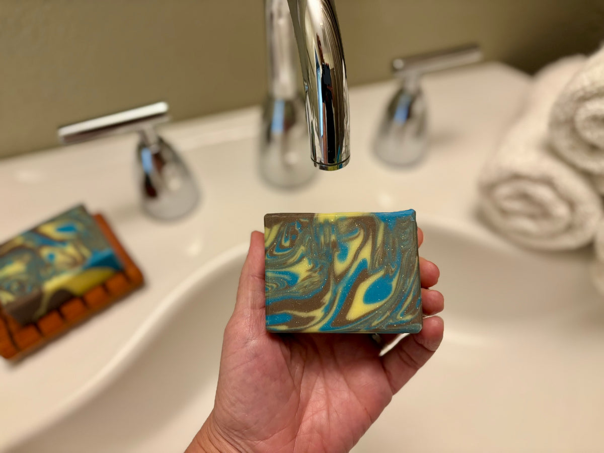 A hand holds a rectangular bar of soap with a blue, brown, and yellow marbled pattern under a chrome faucet in a bathroom setting.
