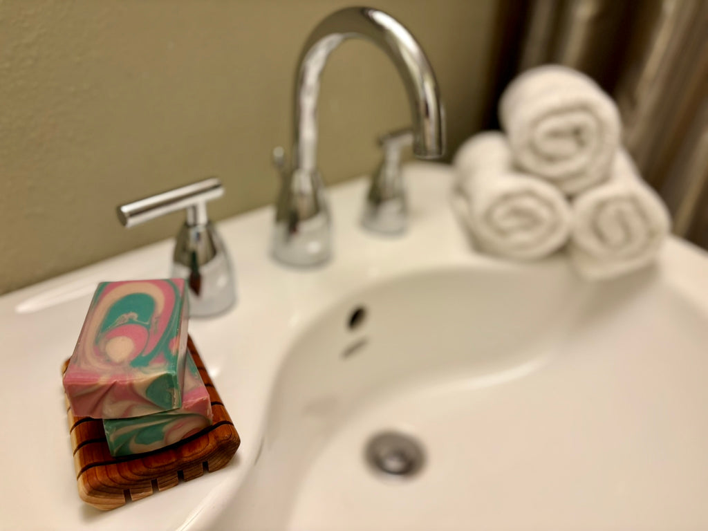 A close-up of a bathroom sink with a chrome faucet, showing a stack of marbled soap bars in pink, green, and teal on a wooden soap dish, with rolled white towels blurred in the background.