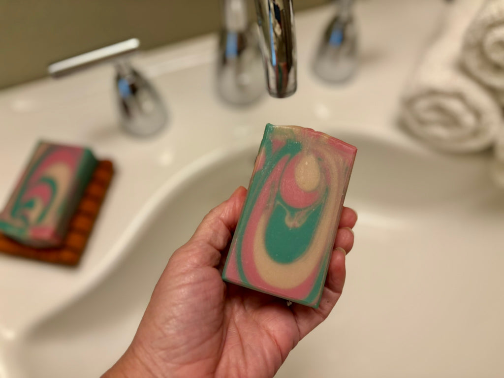 A hand holds a rectangular bar of marbled soap with swirls of pink, teal green, and cream, positioned in front of a white bathroom sink with a chrome faucet.