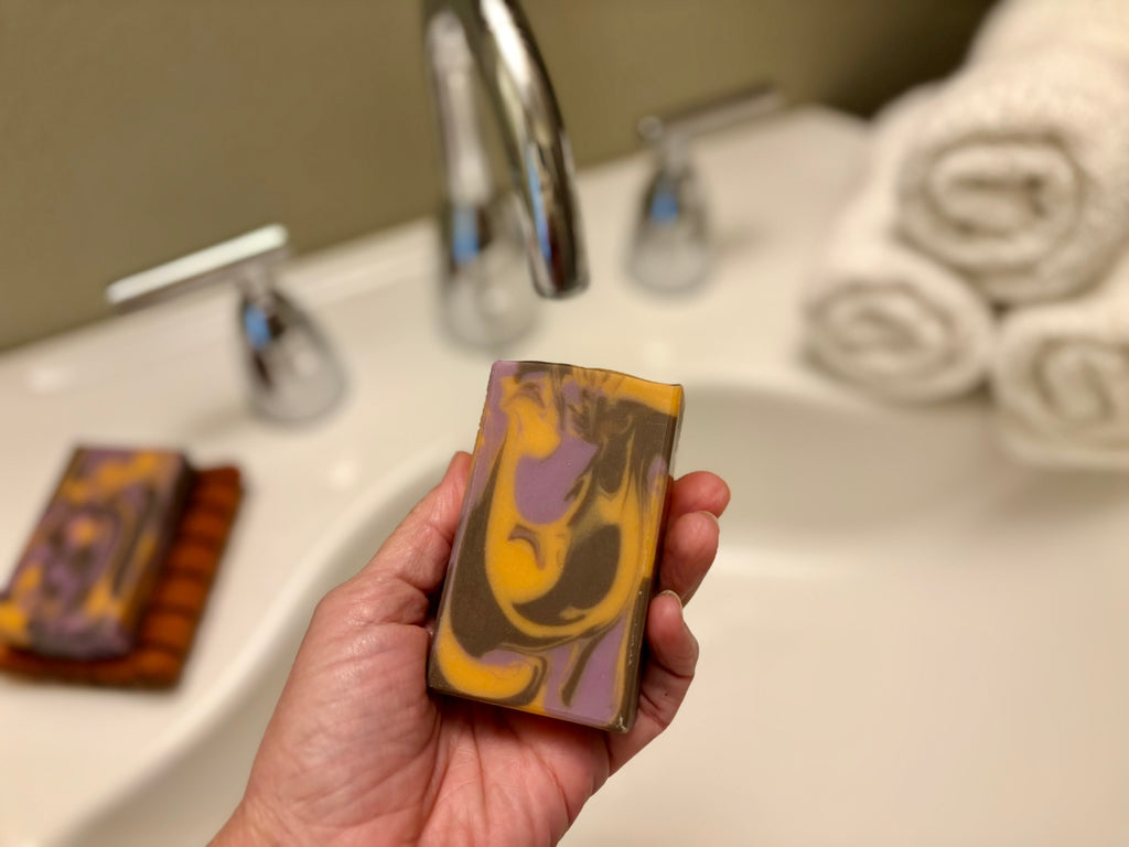 A hand holds a rectangular bar of soap with a marbled swirl pattern in front of a blurred bathroom sink and faucet.