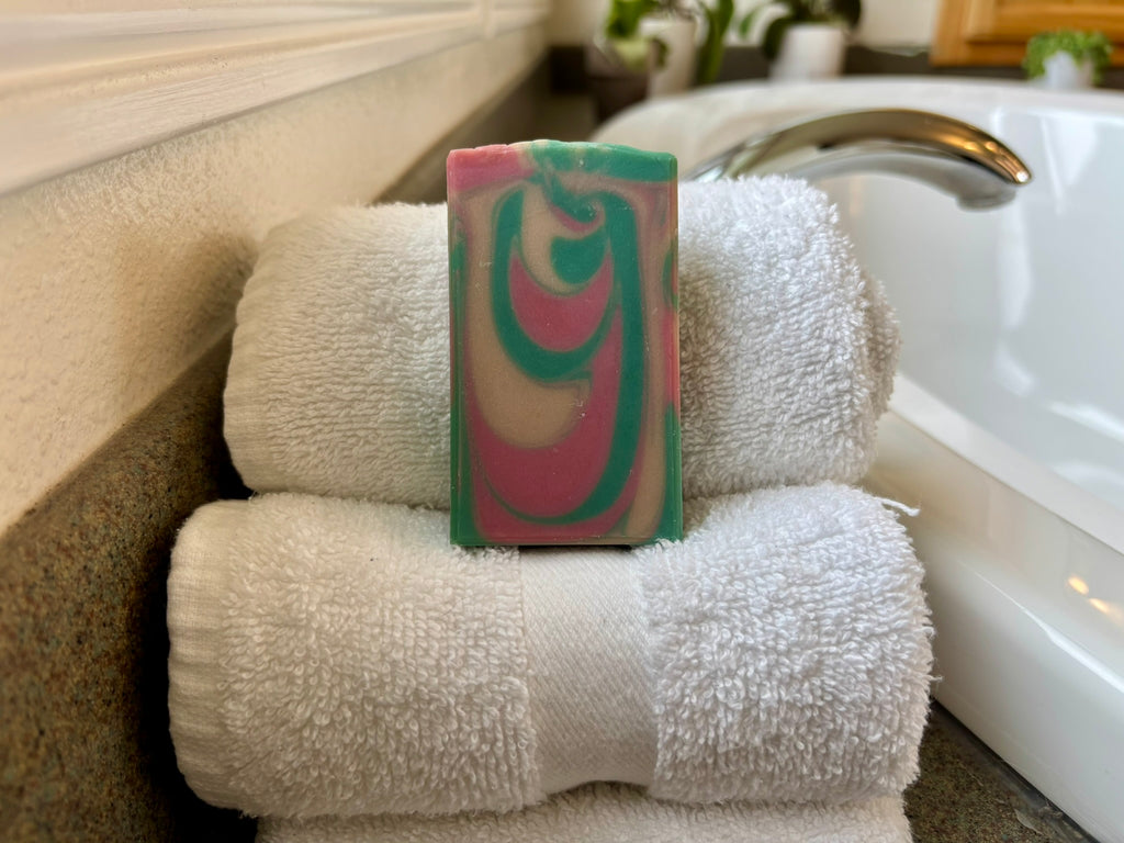 A rectangular handmade soap bar with a pink, green, and white swirl pattern, displayed on top of two folded white towels next to a bathtub faucet.