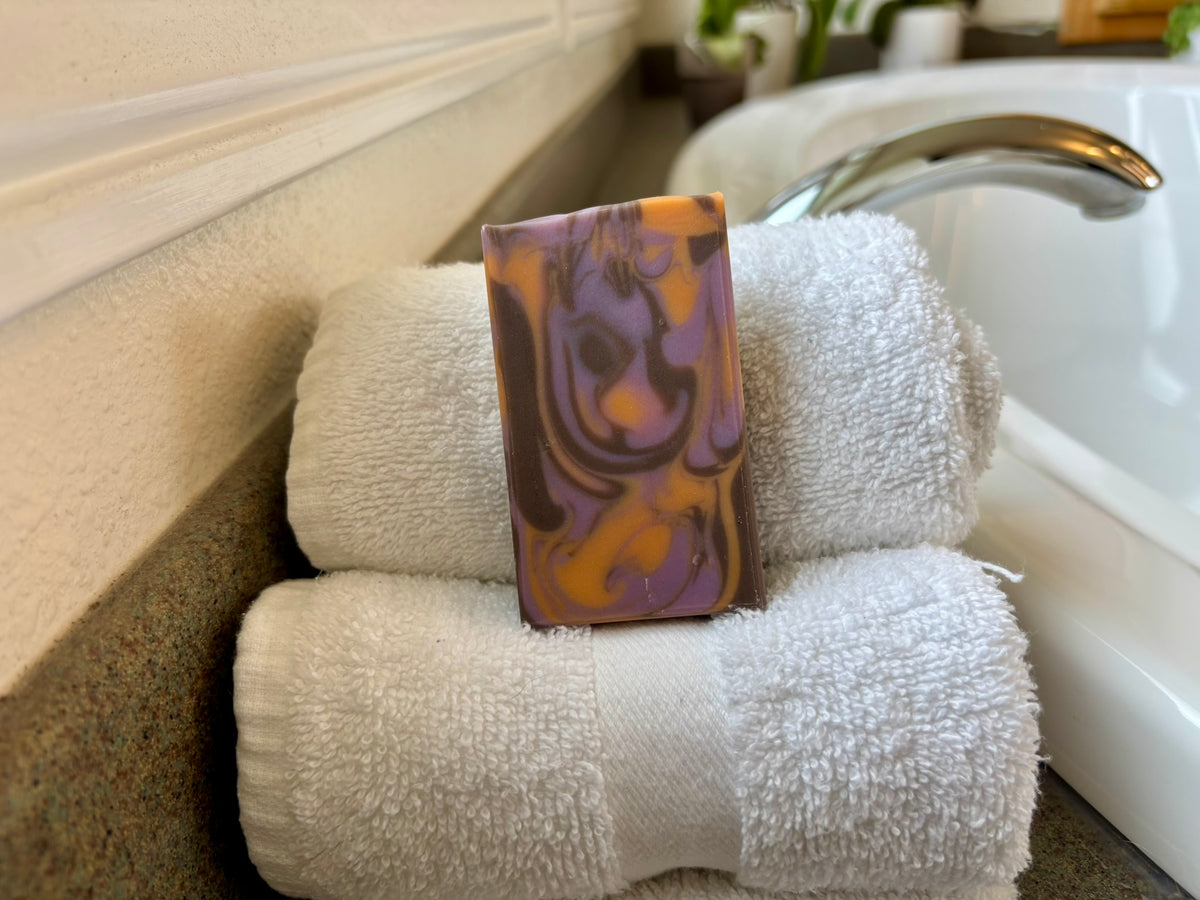 A rectangular bar of marbled soap with swirls of purple, orange, and brown stands upright on top of two folded white towels beside a bathtub faucet.