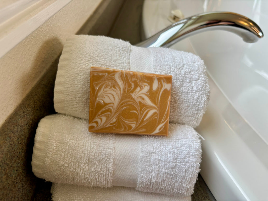 A rectangular bar of marbled tan and cream soap rests on top of two folded white towels beside a chrome faucet and white sink.