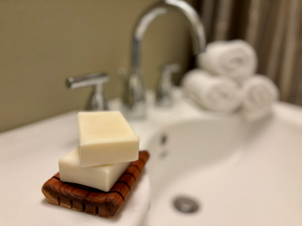 Two stacked rectangular bars of soap on a wooden soap dish, placed on a white bathroom sink with a blurred faucet and rolled towels in the background.