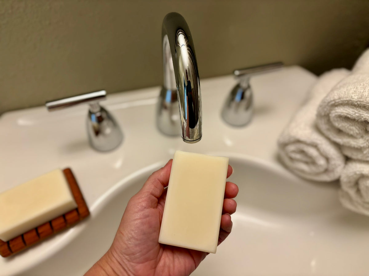 A hand holding a rectangular bar of creamy off-white soap in front of a bathroom sink with a chrome faucet, with a second soap bar on a wooden stand to the left and folded white towels on the right.