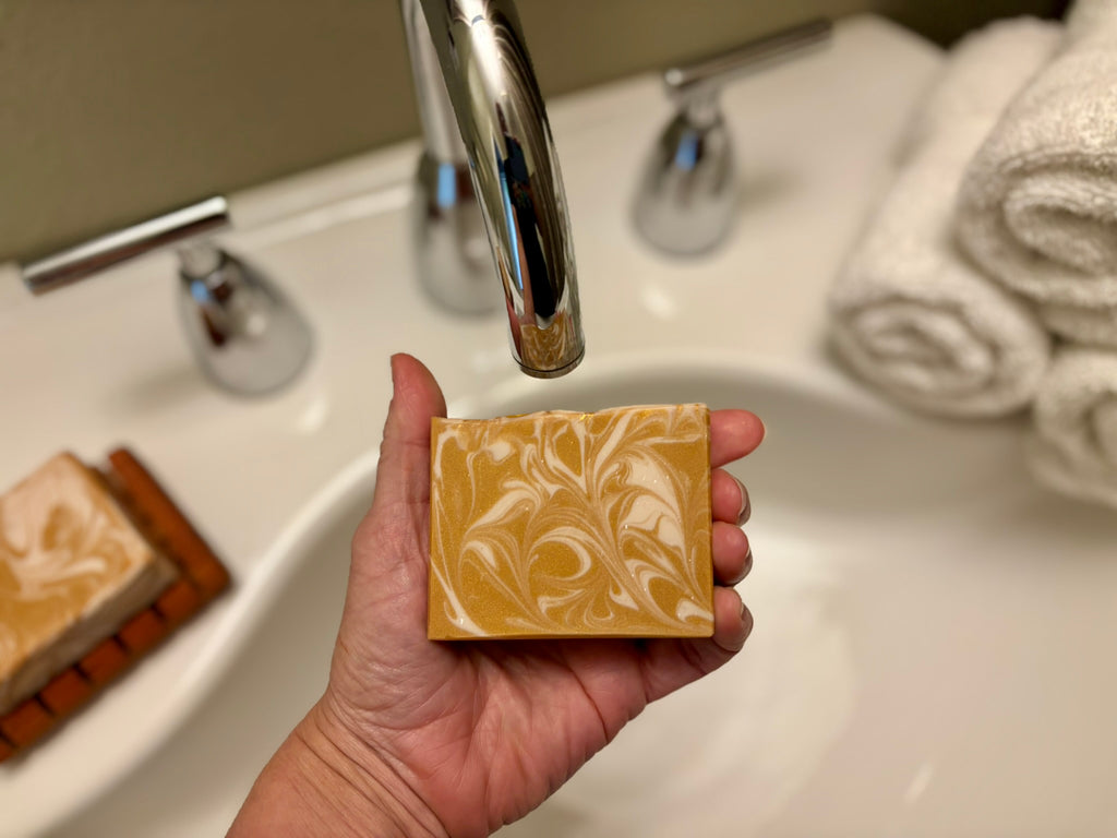 A hand holds a rectangular bar of marbled yellow and white soap under a chrome faucet in a bathroom sink, with rolled towels and another soap bar visible in the background.