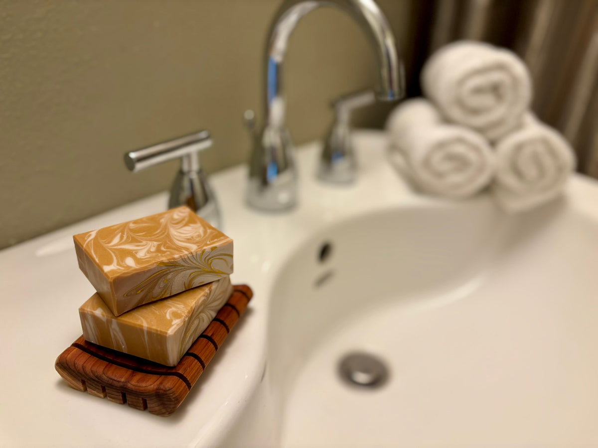 Two stacked rectangular bars of marbled soap in warm beige and tan tones, resting on a dark wooden soap dish on a white bathroom sink. A chrome faucet, white towels, and a beige wall are softly blurred in the background.