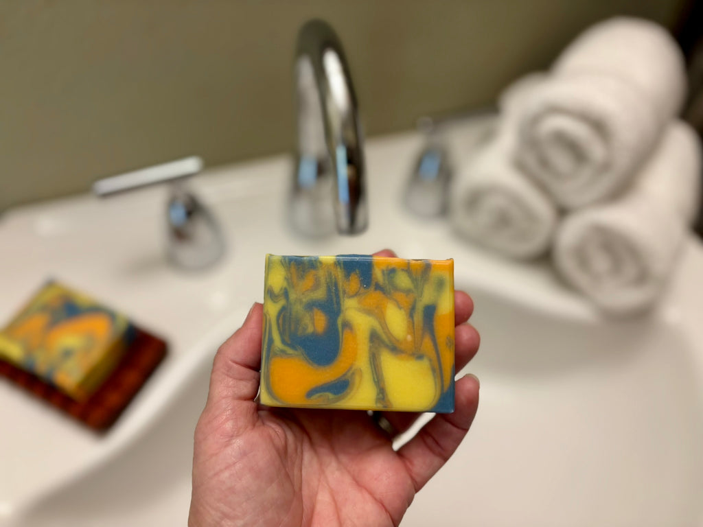 A hand holds a rectangular bar of soap with a marbled yellow, blue, and orange pattern in front of a blurred bathroom sink background with rolled towels.