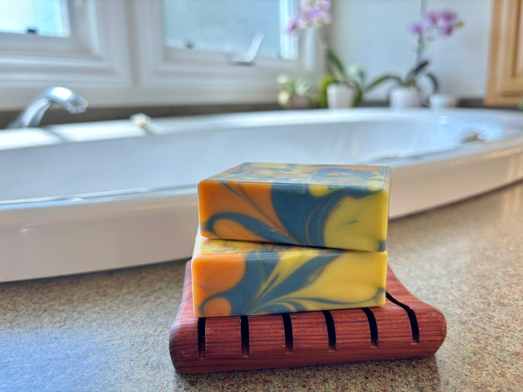 Two stacked rectangular bars of marbled yellow and blue soap on a wooden soap dish, placed on a bathroom countertop with a blurred bathtub and window in the background.