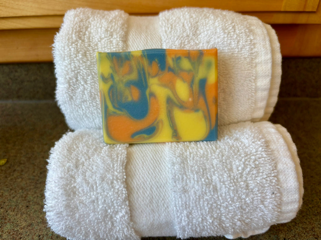 A rectangular bar of marbled soap in yellow, blue, and orange, placed on top of two folded white towels on a countertop.