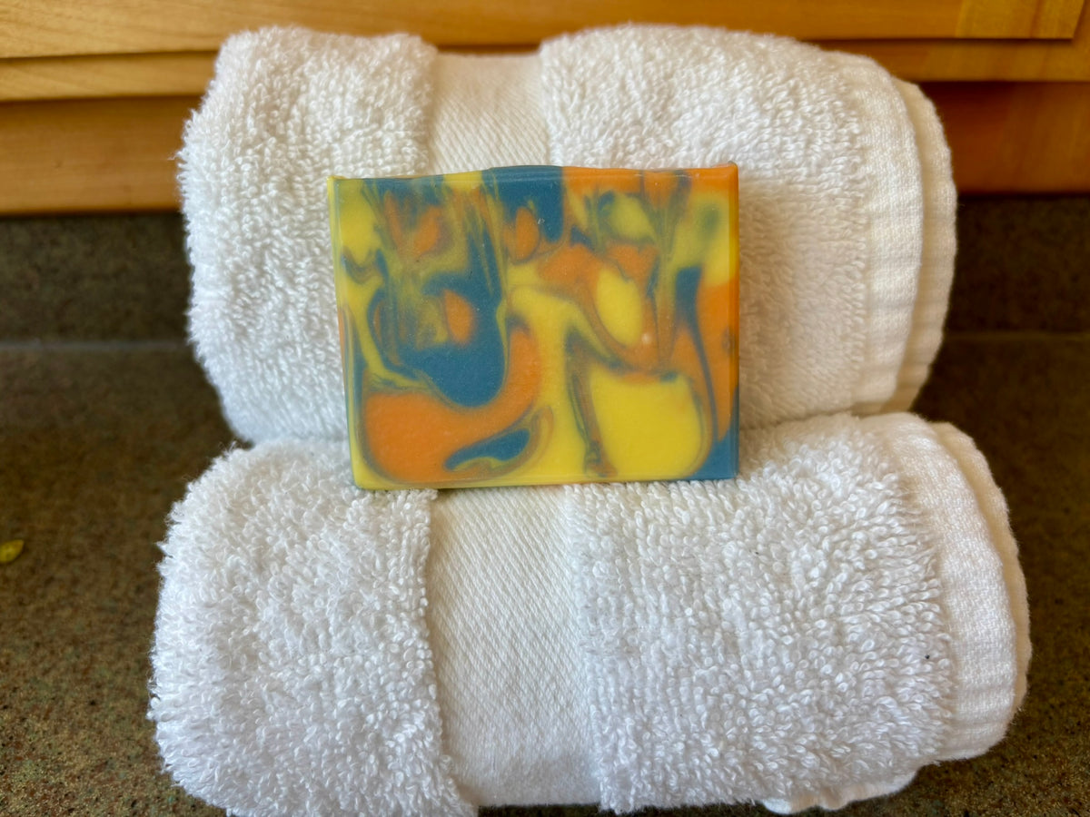 A rectangular bar of marbled soap in yellow, blue, and orange, placed on top of two folded white towels on a countertop.