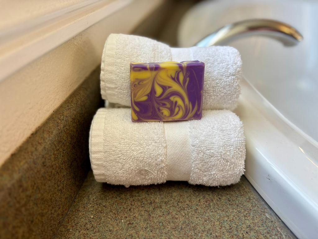 A rectangular bar of soap with a purple and yellow marbled swirl pattern, resting on top of two neatly rolled white towels on a bathroom counter next to a sink.