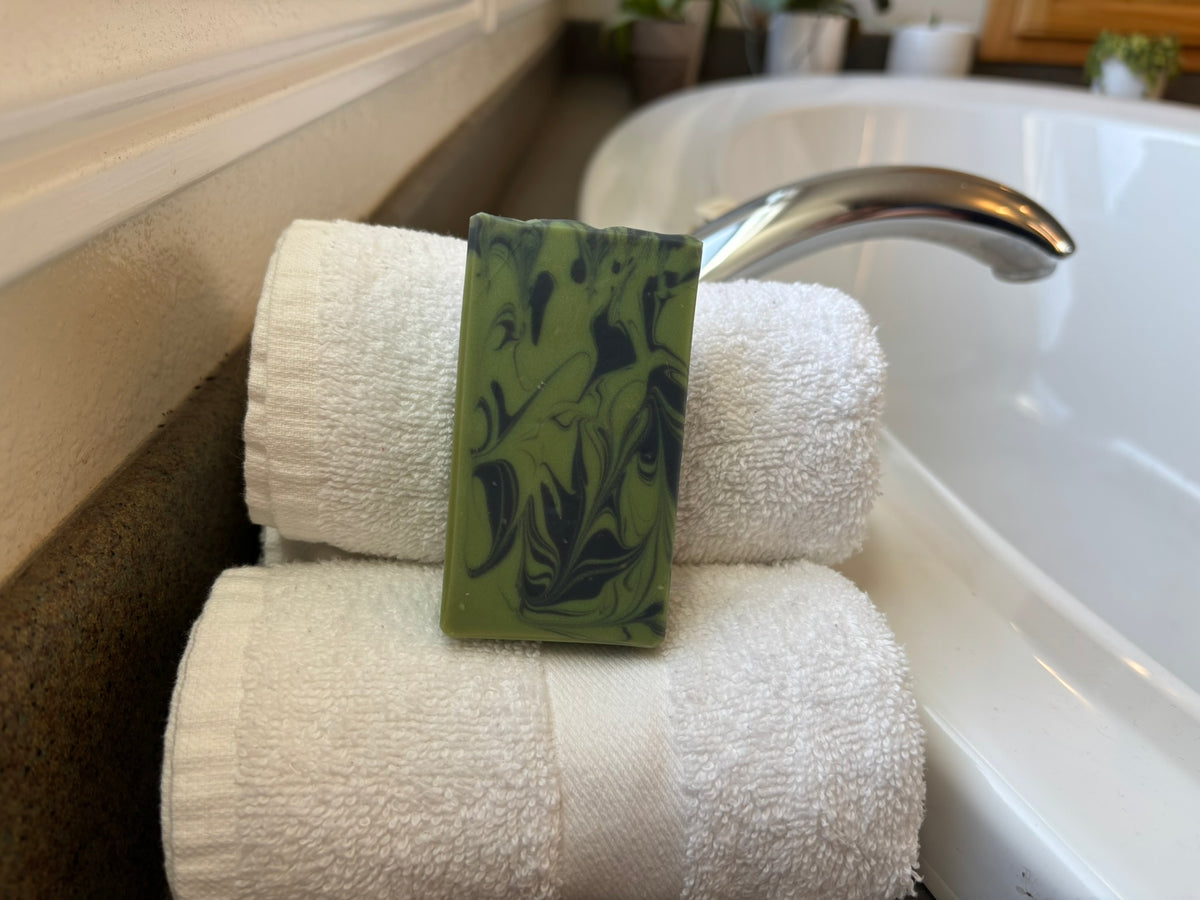A rectangular green soap bar with a marbled pattern rests on top of two rolled white towels beside a bathtub faucet.