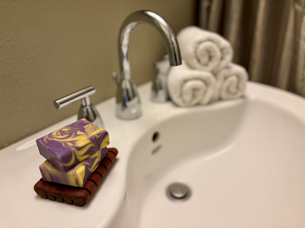 Two stacked rectangular bars of marbled purple and yellow soap on a wooden soap dish, placed on a white bathroom sink next to a chrome faucet with rolled white towels in the background.