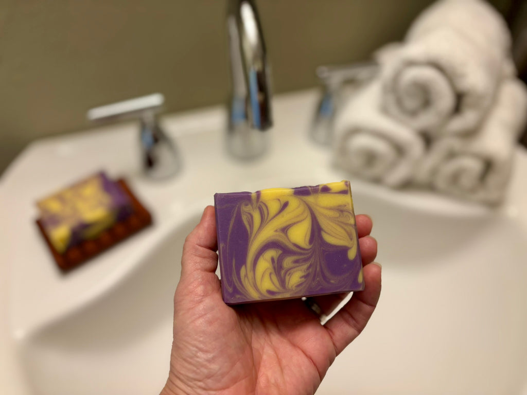 A hand holds a rectangular bar of soap with a purple and yellow marbled swirl pattern in front of a blurred bathroom sink background.
