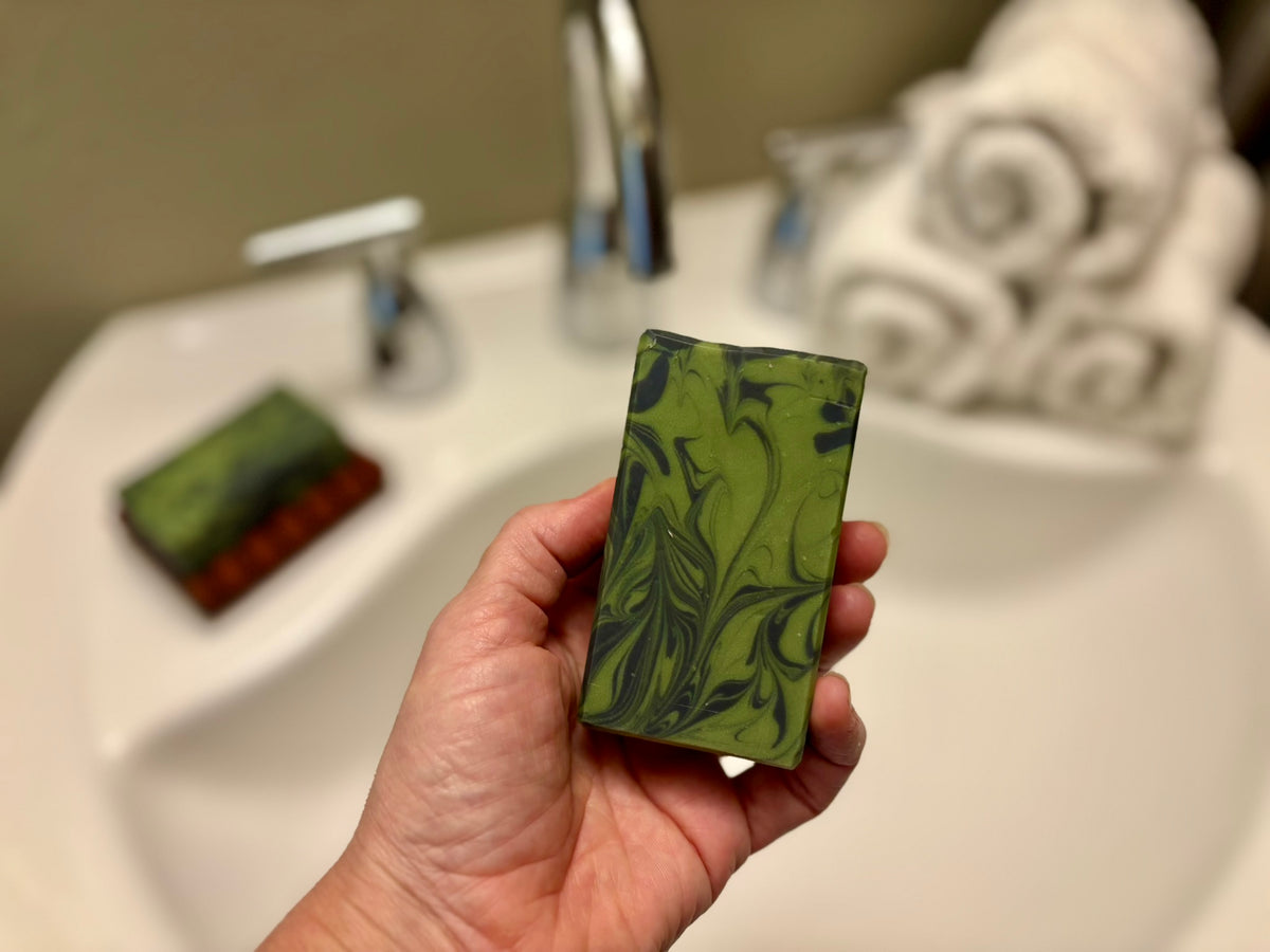 A hand holds a rectangular bar of green marbled soap in front of a blurred bathroom sink and faucet.