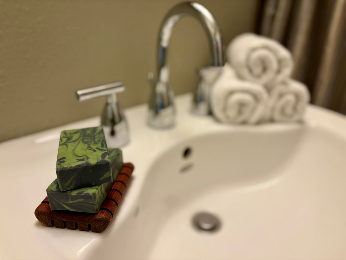 Two stacked rectangular bars of soap with a green and gray marbled pattern, resting on a wooden soap dish on a white bathroom sink next to a chrome faucet.