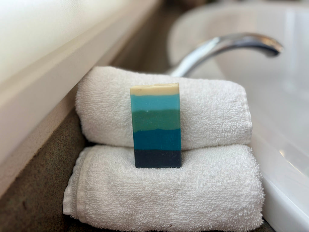 A rectangular bar of soap with horizontal stripes of cream, green, teal, and dark navy rests on top of two folded white towels beside a sink and faucet.