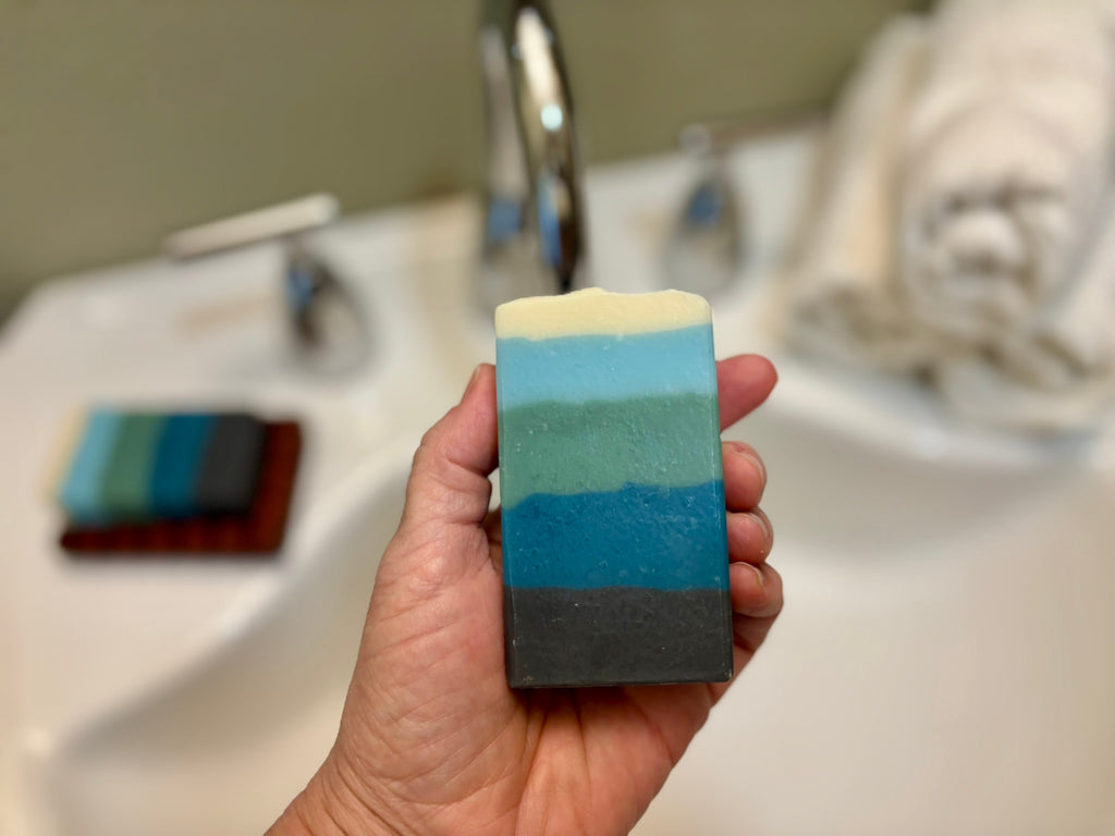 A hand holds a color bar of soap in front of a sink with a chrome faucet.