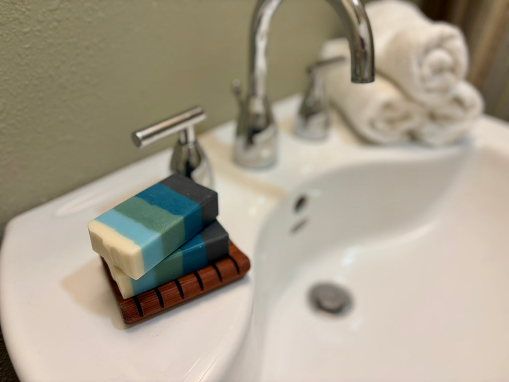 Two striped bar soaps in shades of cream, light blue, teal, and dark gray, resting on a wooden soap dish on a white bathroom sink next to a chrome faucet.