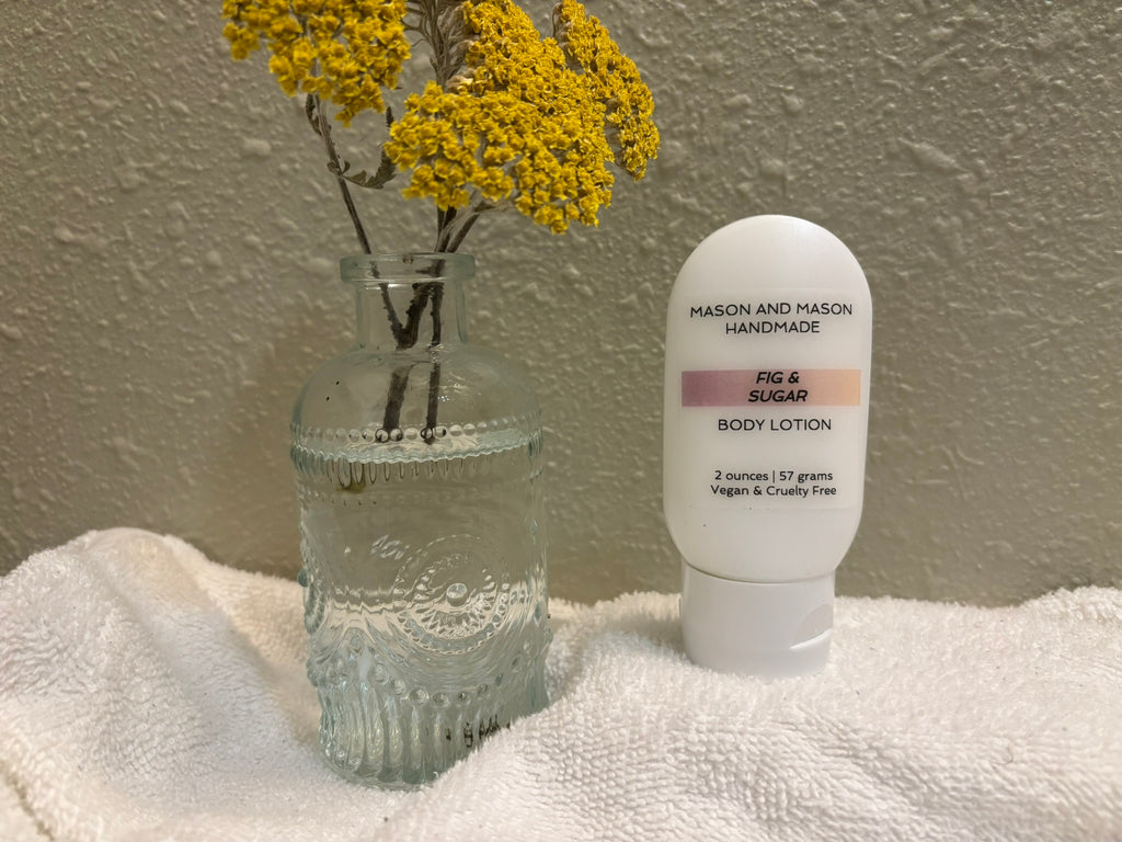 Two ounce body lotion bottle labeled 'Mason and Mason Handmade' next to a clear glass vase with yellow flowers on a white surface.