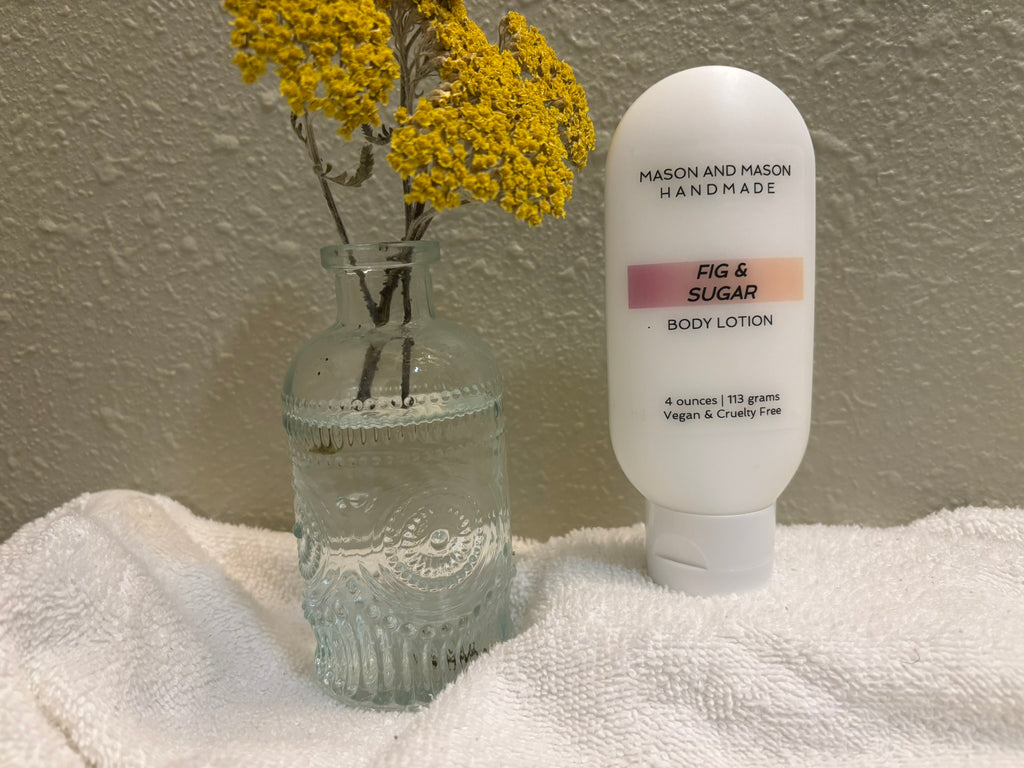 Four ounce body lotion bottle labeled 'Mason and Mason Handmade' next to a vase with yellow flowers on a white surface.