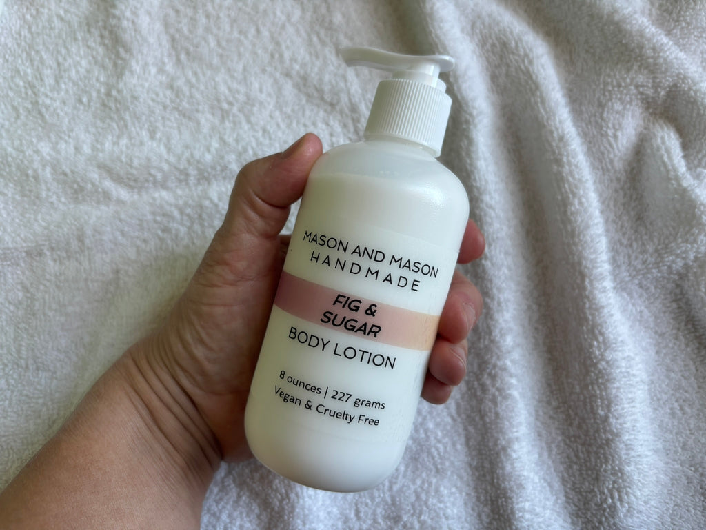 Hand holding a bottle of Mason and Mason Handmade eight ounce Fig & Sugar Body Lotion, with a pump, on a textured white background