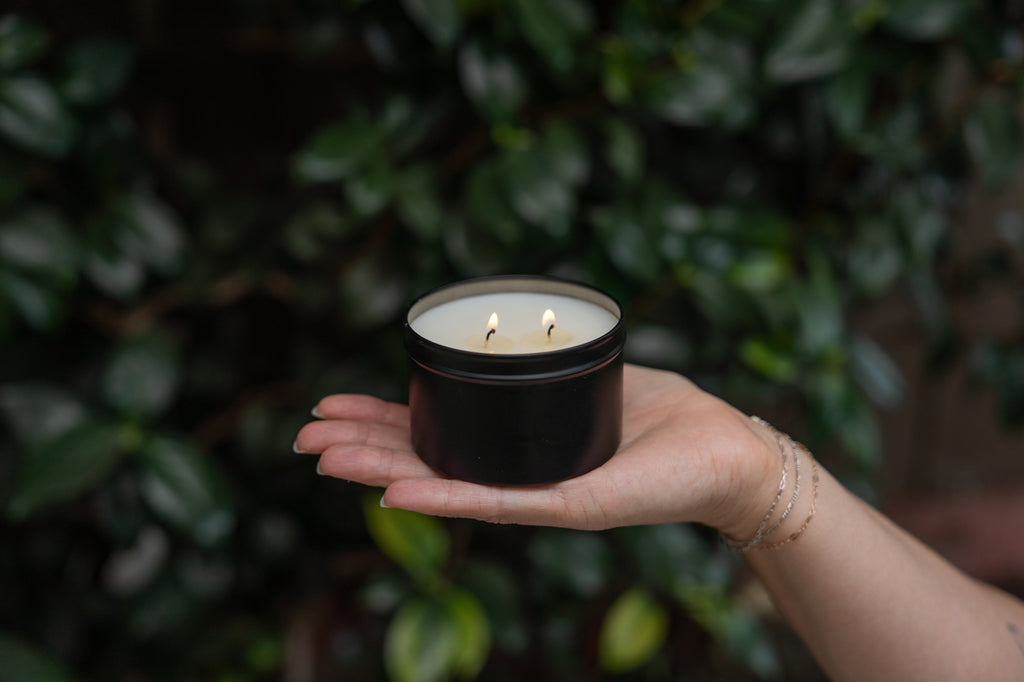 A black metal tin candle with two lit wicks, held in a person's hand against a blurred background of green foliage.