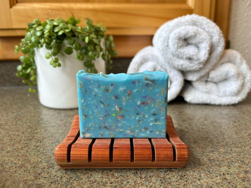 Blue speckled soap on a wooden soap dish with towels and a plant in the background.