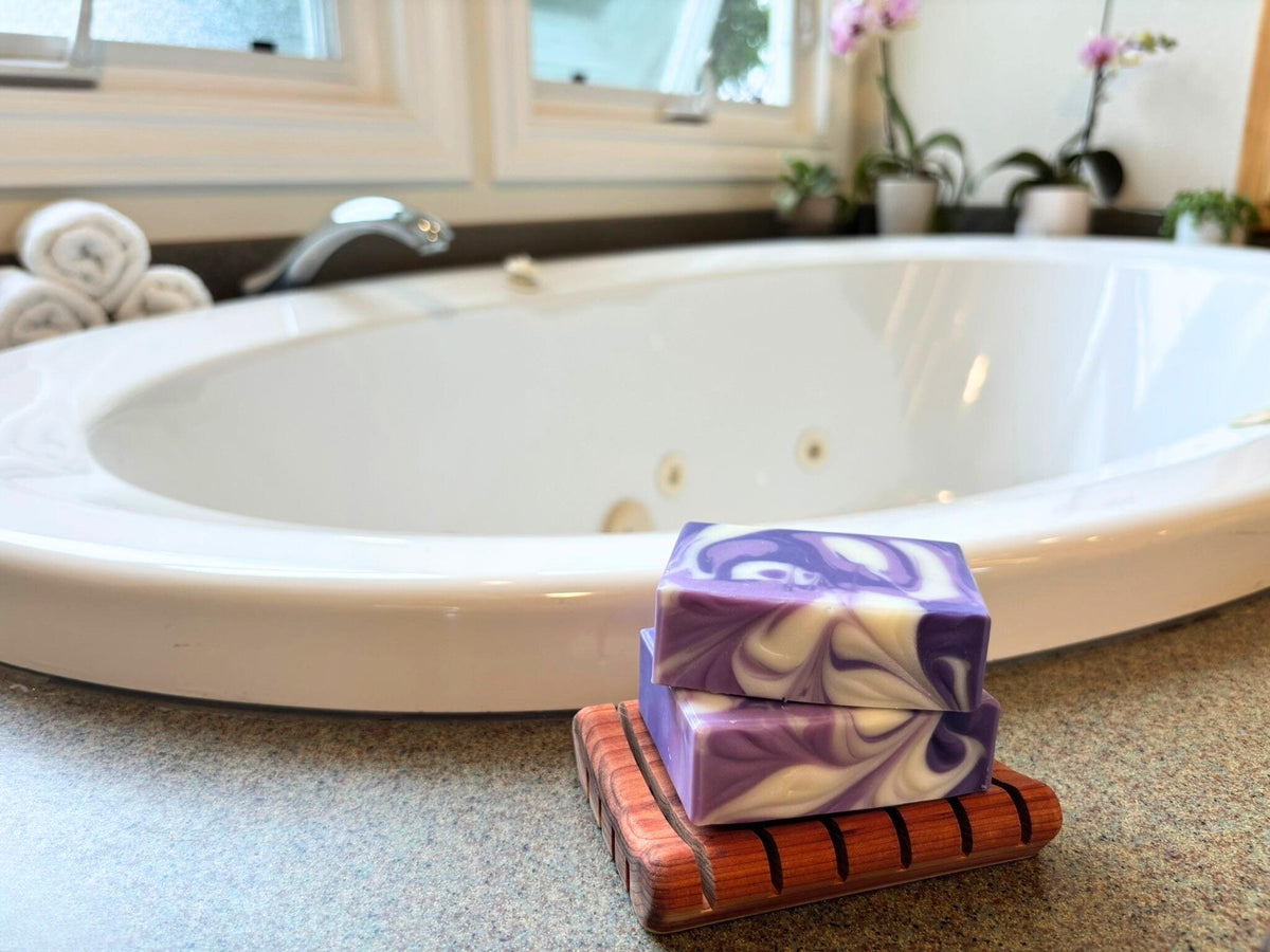 Stack of purple and white swirled soap bars on a wooden soap dish in front of a bathtub.