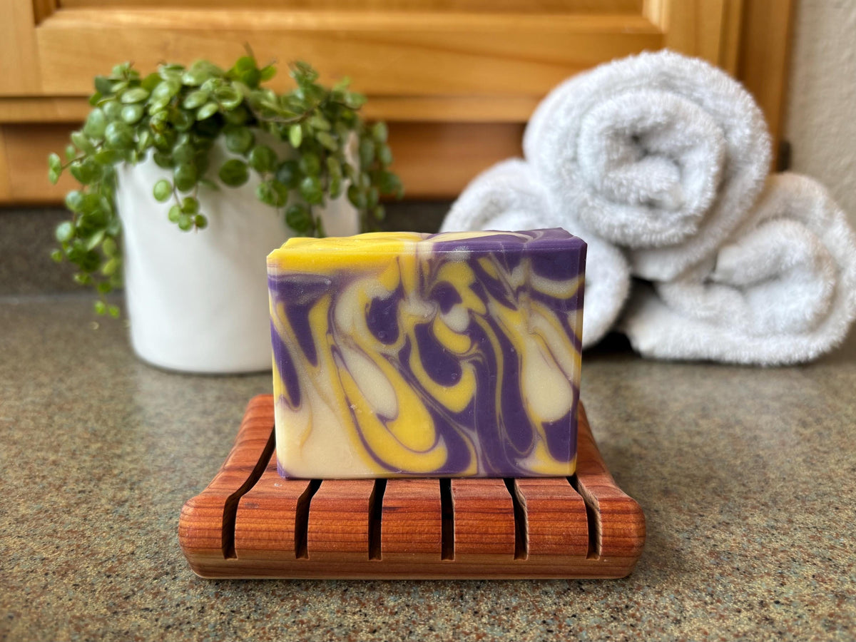 Swirled purple and yellow soap on a wooden soap dish with towels and a plant in the background.