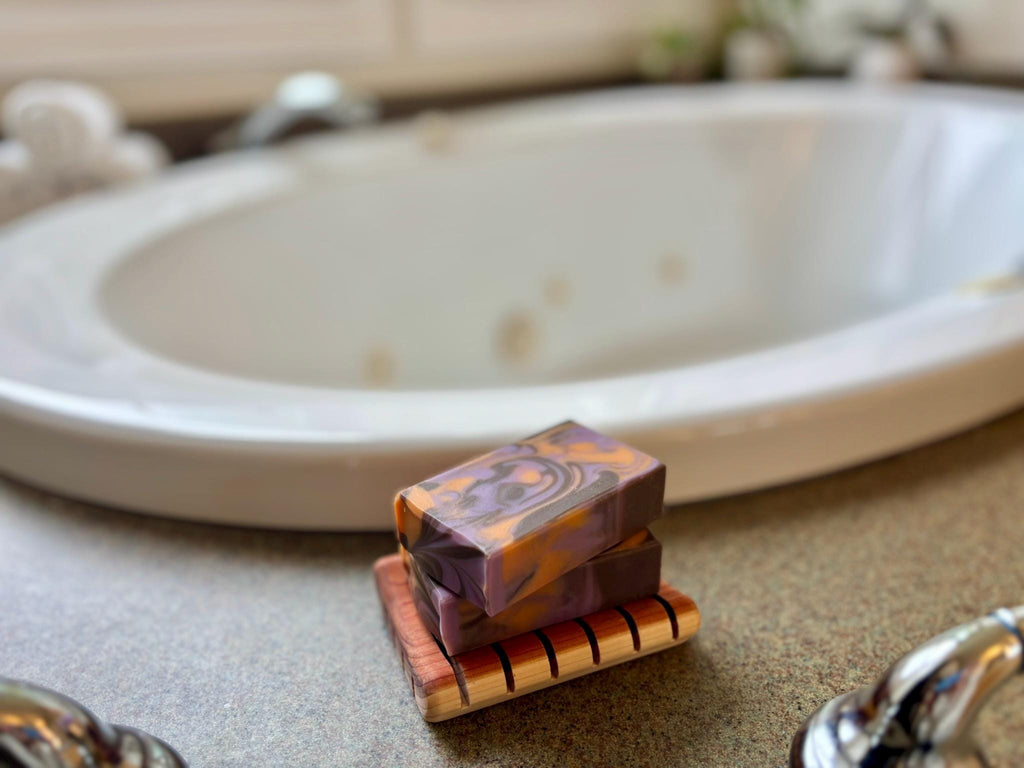 Two soap bars on a wooden soap dish with a blurred bathtub in the background.
