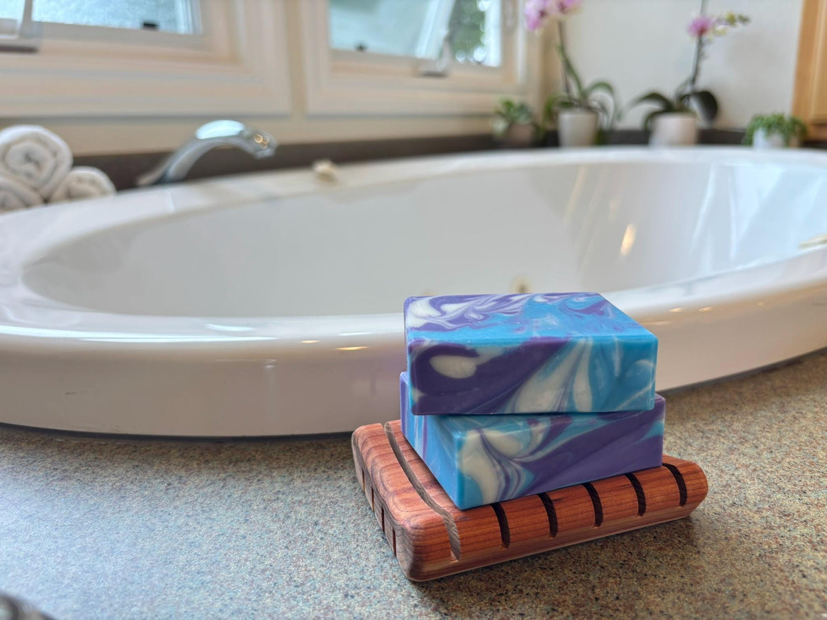 Two bars of colorful soap stacked on a wooden soap dish with a bathtub in the background.