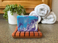 Marbled blue and purple soap on a wooden soap dish with towels and a plant in the background.