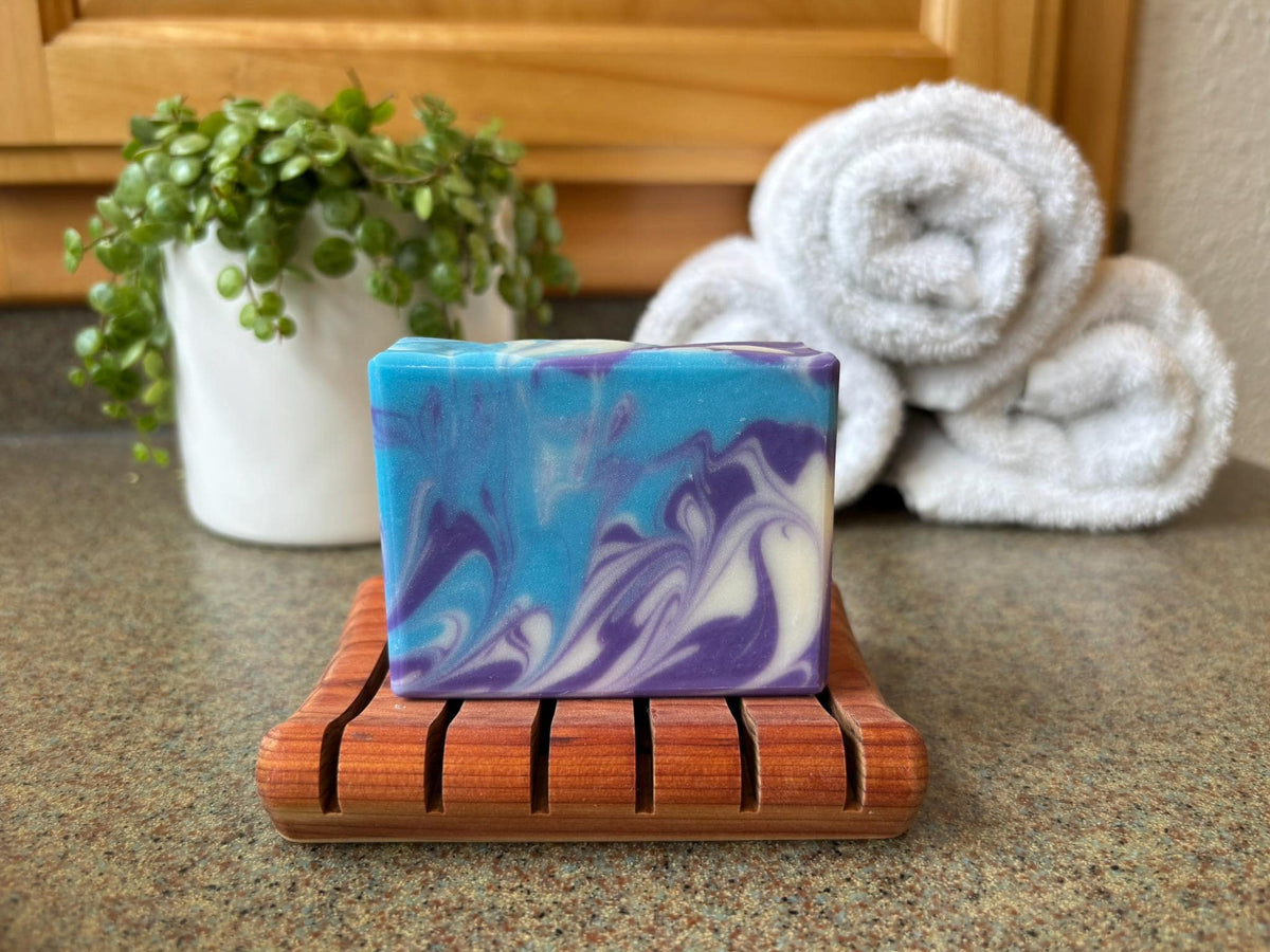 Marbled blue and purple soap on a wooden soap dish with towels and a plant in the background.